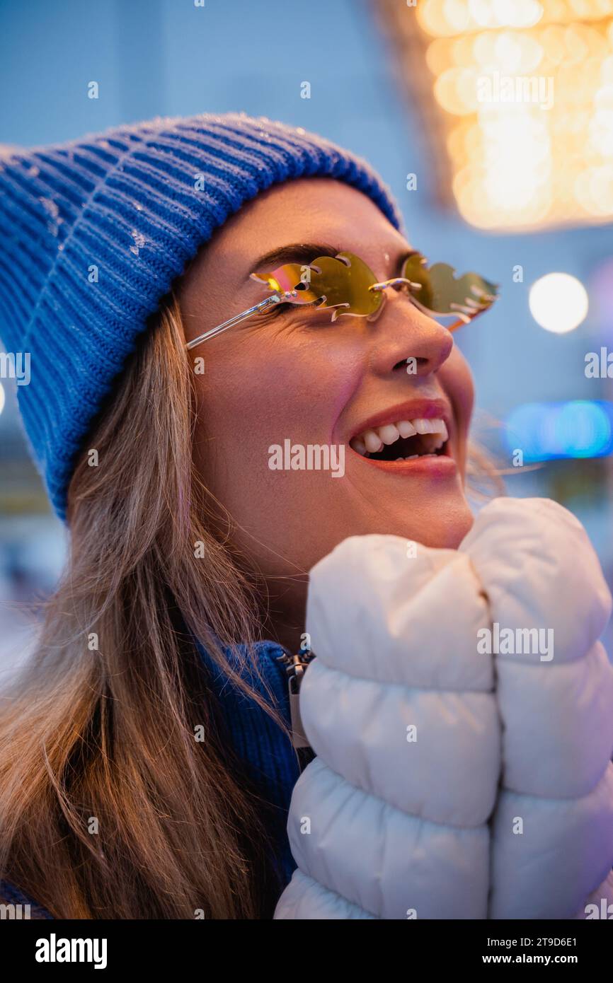 Fröhliche und stilvolle Frau, in warmen Kleidern gekleidet und mit einer Sonnenbrille in Form von Feuerflammen, hat Spaß in einem verschneiten Winterspazierpark. Stockfoto