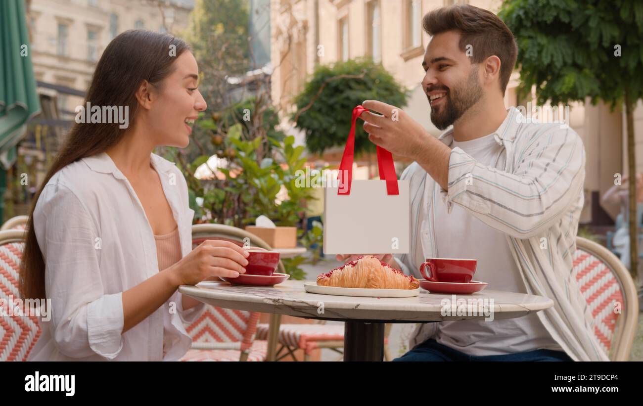 Glückliches europäisches Paar Mann gibt Frau ein Geschenk Überraschung Jubiläum Feier Emotionen Stadt draußen Café zusammen zarte Liebe Gefühle lächelnd Stockfoto