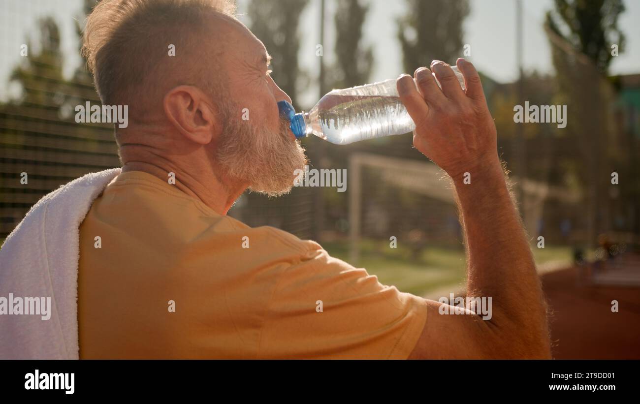 Rückansicht Alter männlicher Sportler Athlet Walking Stadion draußen trinken kaltes Wasser aus Sportflasche Kaukasischer Mann Rentner trinken Löschdurst Stockfoto
