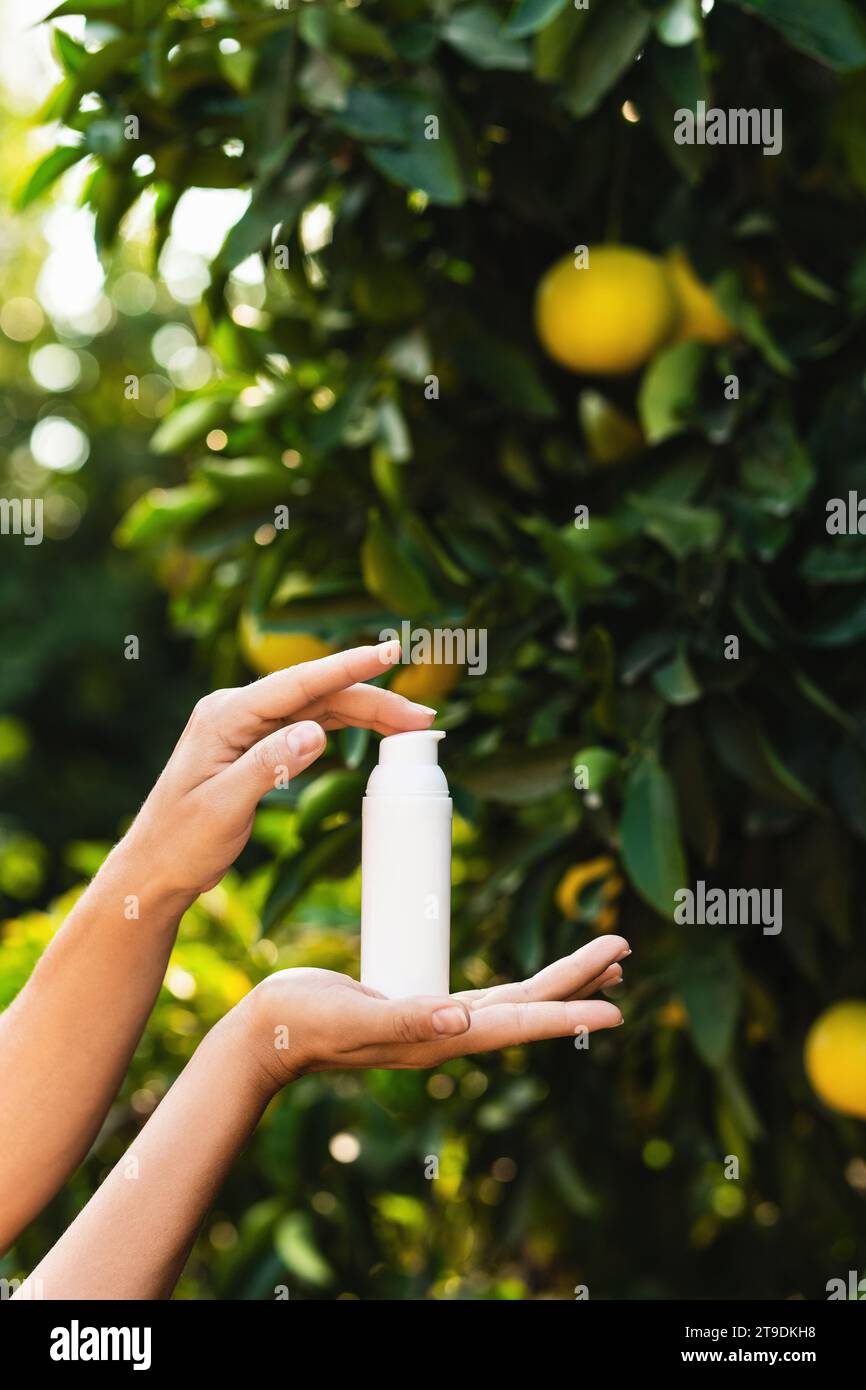 Frau hält eine weiße Flasche Hautpflegemittel in den Händen auf verschwommenem Zitronenbaumhintergrund. Stockfoto