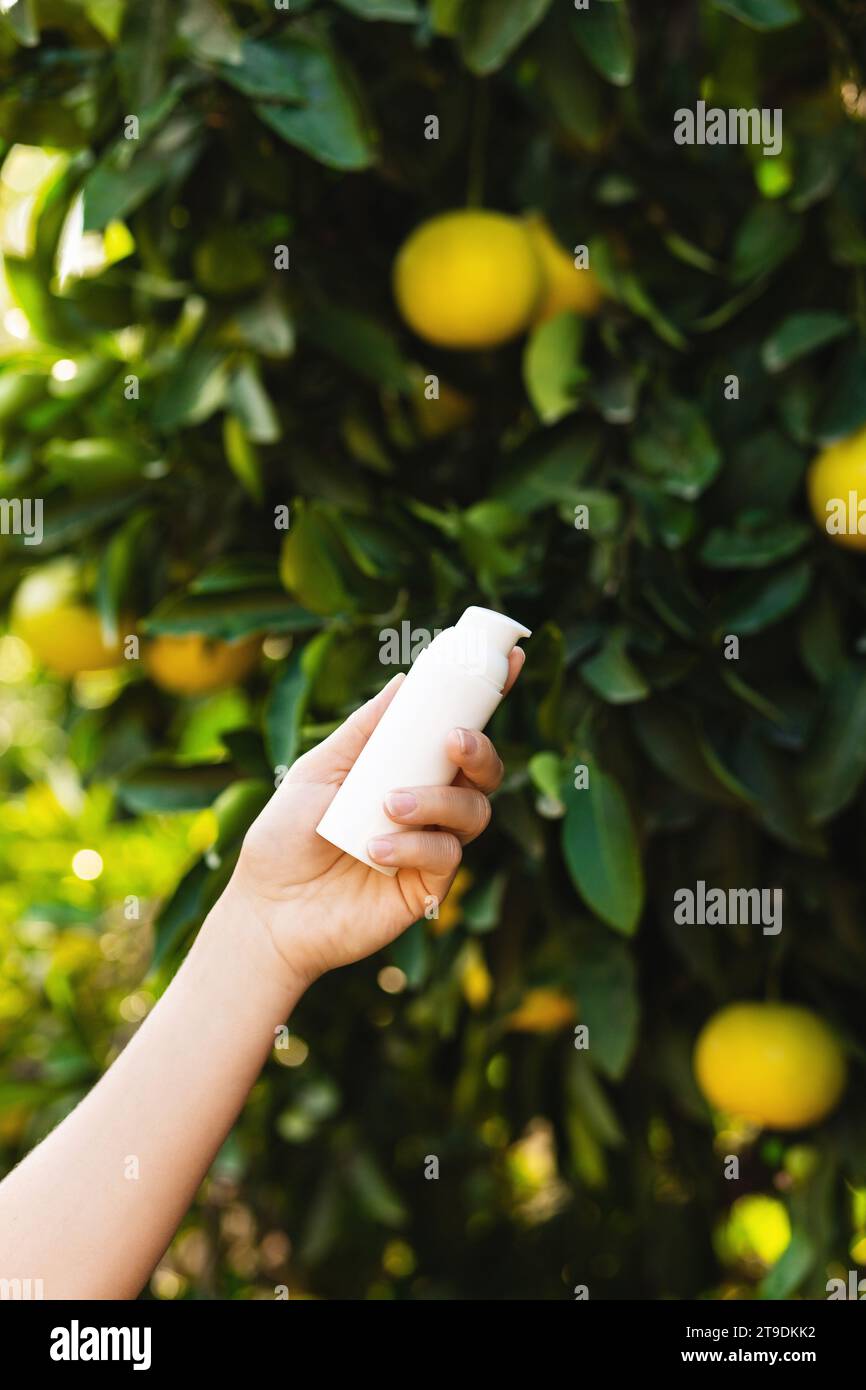 Frau hält eine weiße Flasche Hautpflegemittel in der Hand auf verschwommenem Zitronenbaumhintergrund. Stockfoto