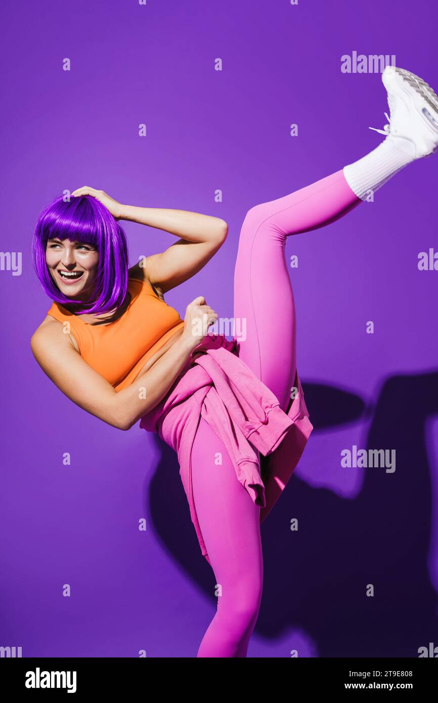 Sorglose junge Tänzerin in farbenfroher Sportkleidung, die vor purpurrotem Hintergrund auftritt Stockfoto