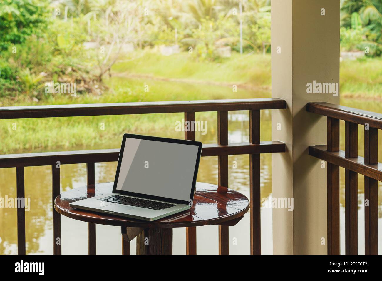 Laptop mit leerem Bildschirm auf einem kleinen Holztisch auf der Terrasse einer Hütte in einer ruhigen ländlichen Gegend. Stockfoto