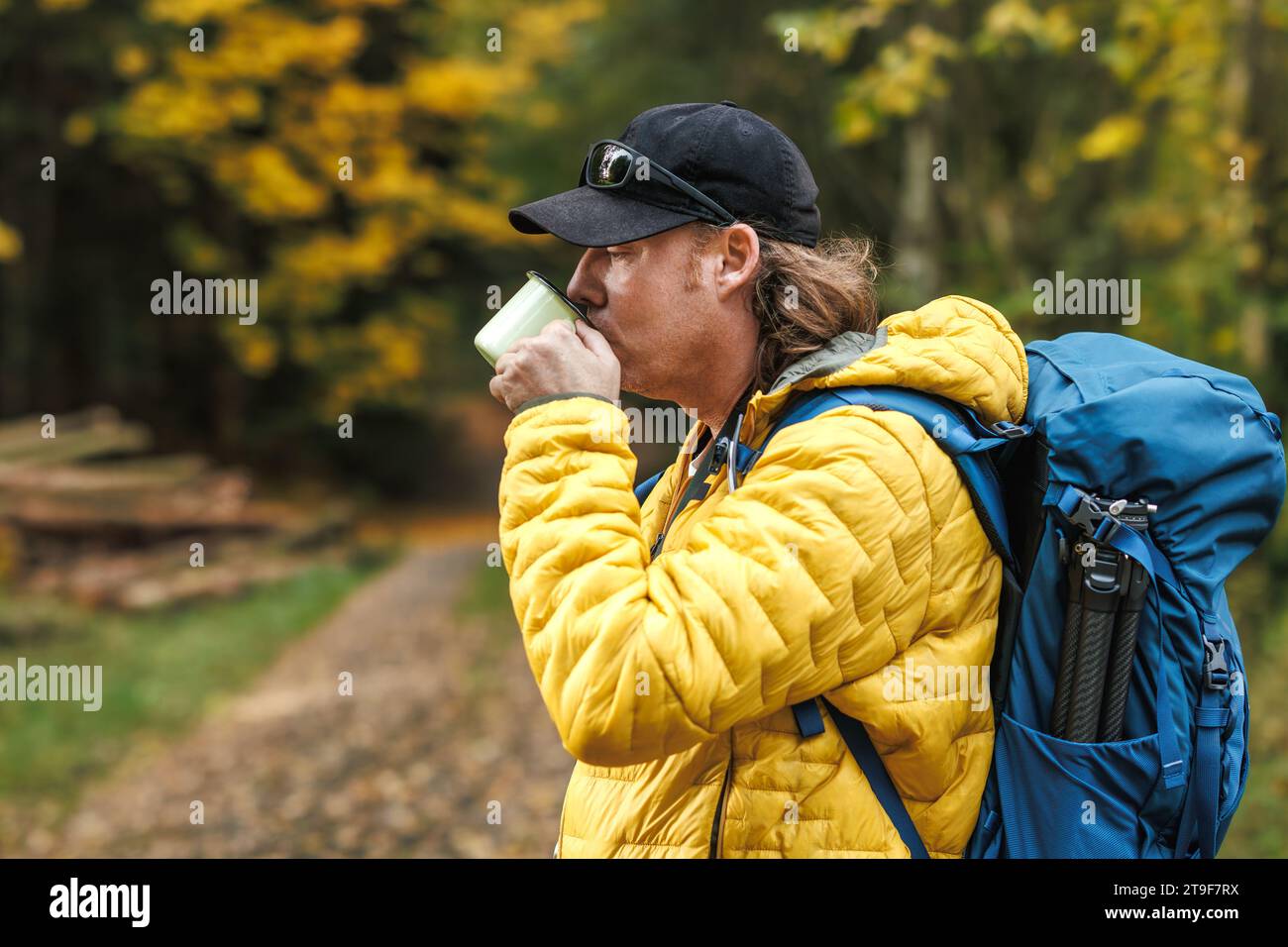 Wanderer mit Rucksack und gelber Jacke trinkt heißen Tee aus Reisebecher im Herbstwald. Erfrischung und Erholung bei Wanderungen im Freien Stockfoto