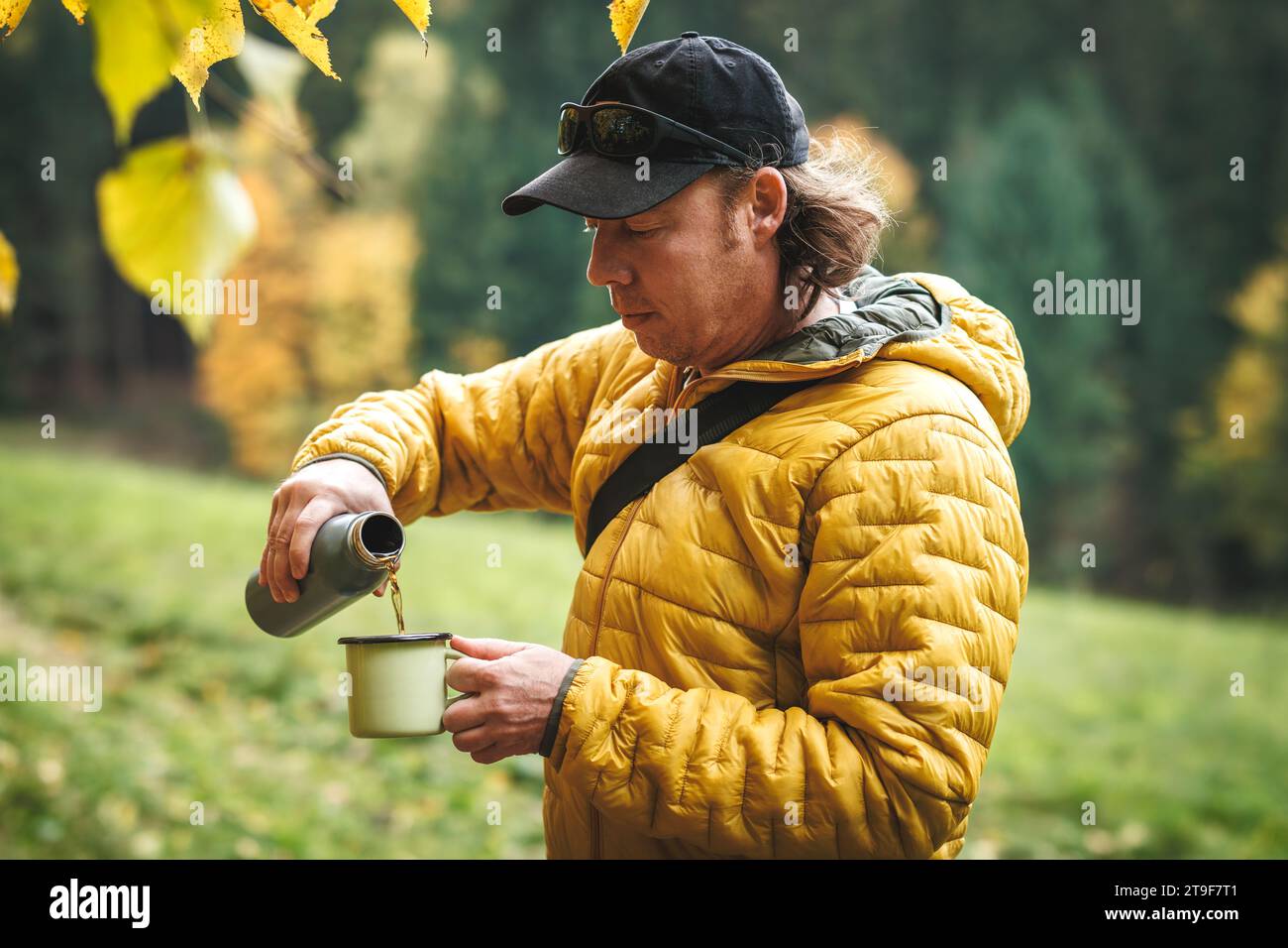 Ein Wanderer mit gelber Jacke gießt heißes Getränk aus der Thermoskanne in einen Reisebecher im Herbstwald. Erholung und Erfrischung bei Wanderungen im Freien Stockfoto
