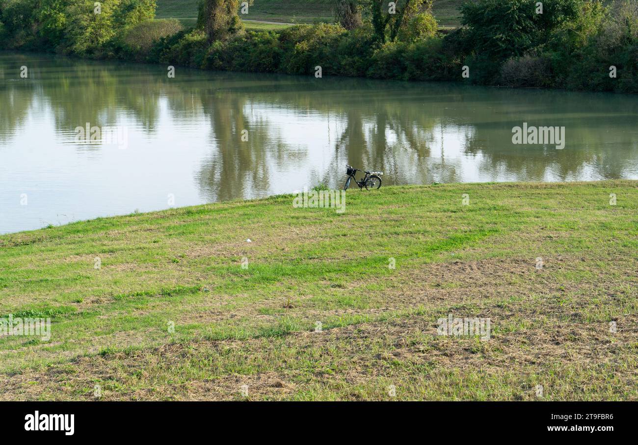 Ein Fahrrad, das am Ufer eines Flusses während einer ökologischen Natur-orientierten Intinerary geparkt wurde. Grünes Gras und Bäume im Hintergrund tragen zum Ser bei Stockfoto