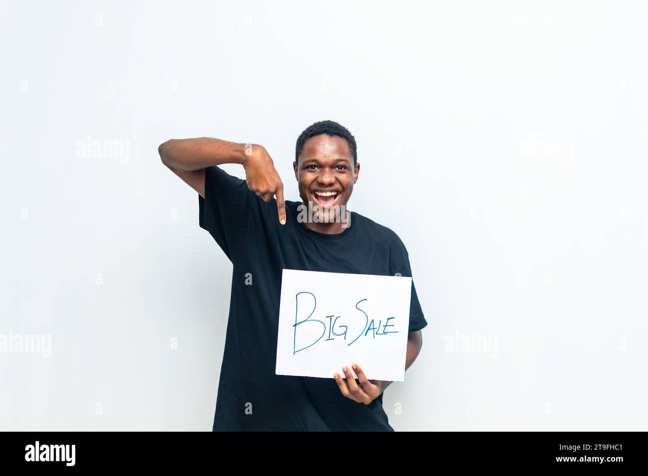 Aufgeregter afrikanischer Mann mit schwarzem Hemd, der auf eine weiße Karte mit Big Sale-Aufschrift zeigt und damit den Abschluss großer Verkäufe ankündigt. Black Friday s Stockfoto