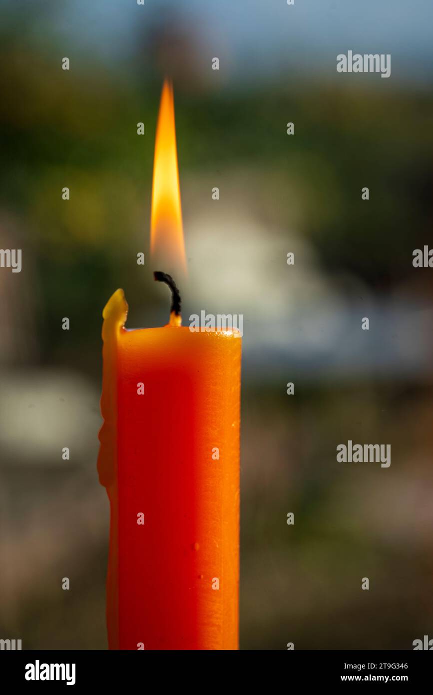 Eine einsame orangene Kerze strahlt ein warmes Leuchten in einem Fenster aus, die Flamme flackert vor einem verschwommenen Hintergrund im Freien. Die sanfte Beleuchtung erzeugt einen SER Stockfoto