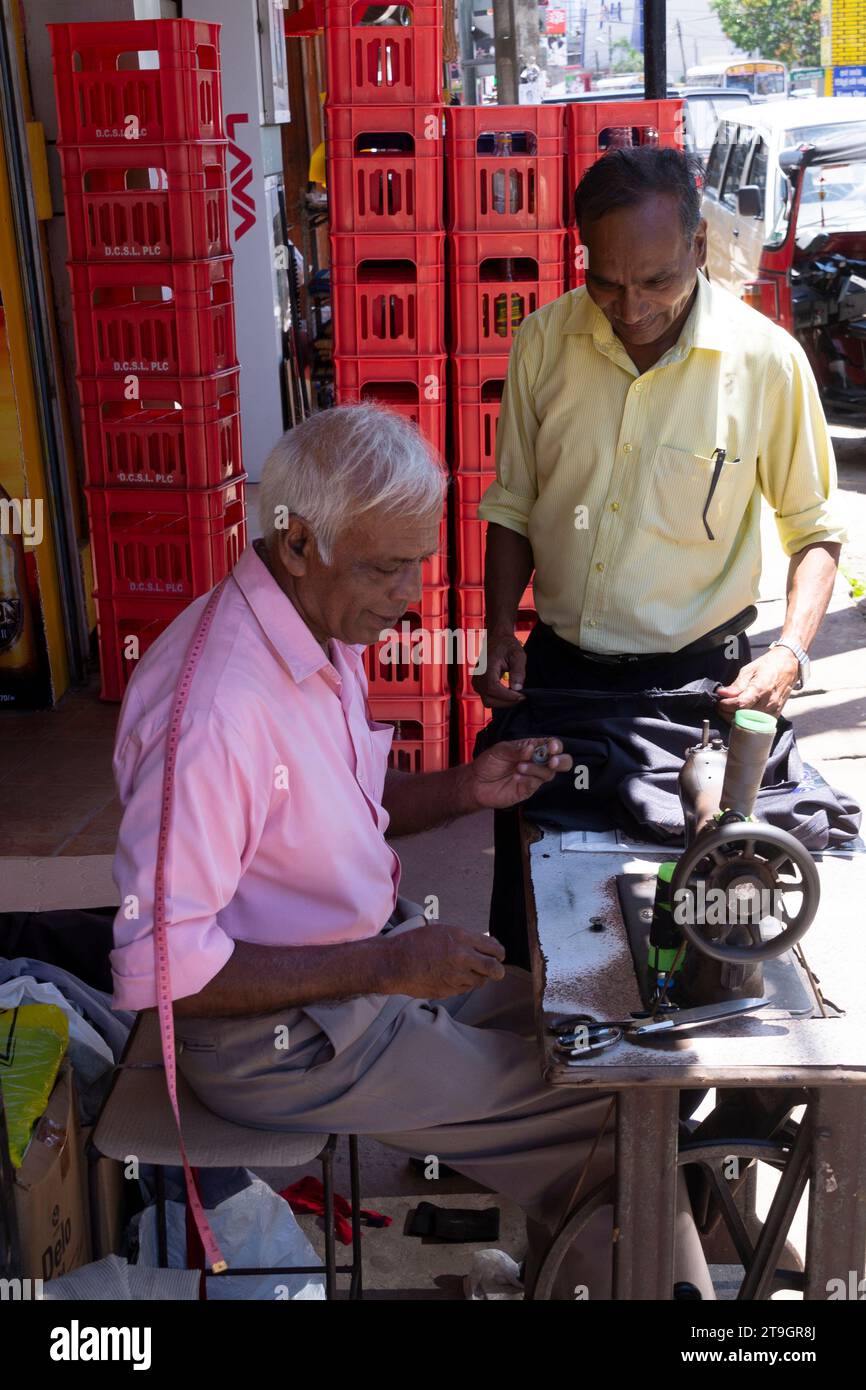 Ein Mann benutzt eine Nähmaschine, während ein anderer vor einem Geschäft in Sri Lanka zusieht Stockfoto