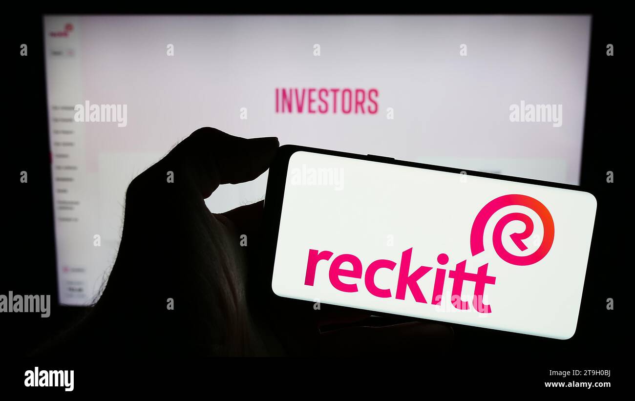 Person, die ein Mobiltelefon mit dem Logo des Konsumgüterunternehmens Reckitt Benckiser Group plc vor der Business-Webseite hält. Schwerpunkt auf dem Display des Telefons. Stockfoto