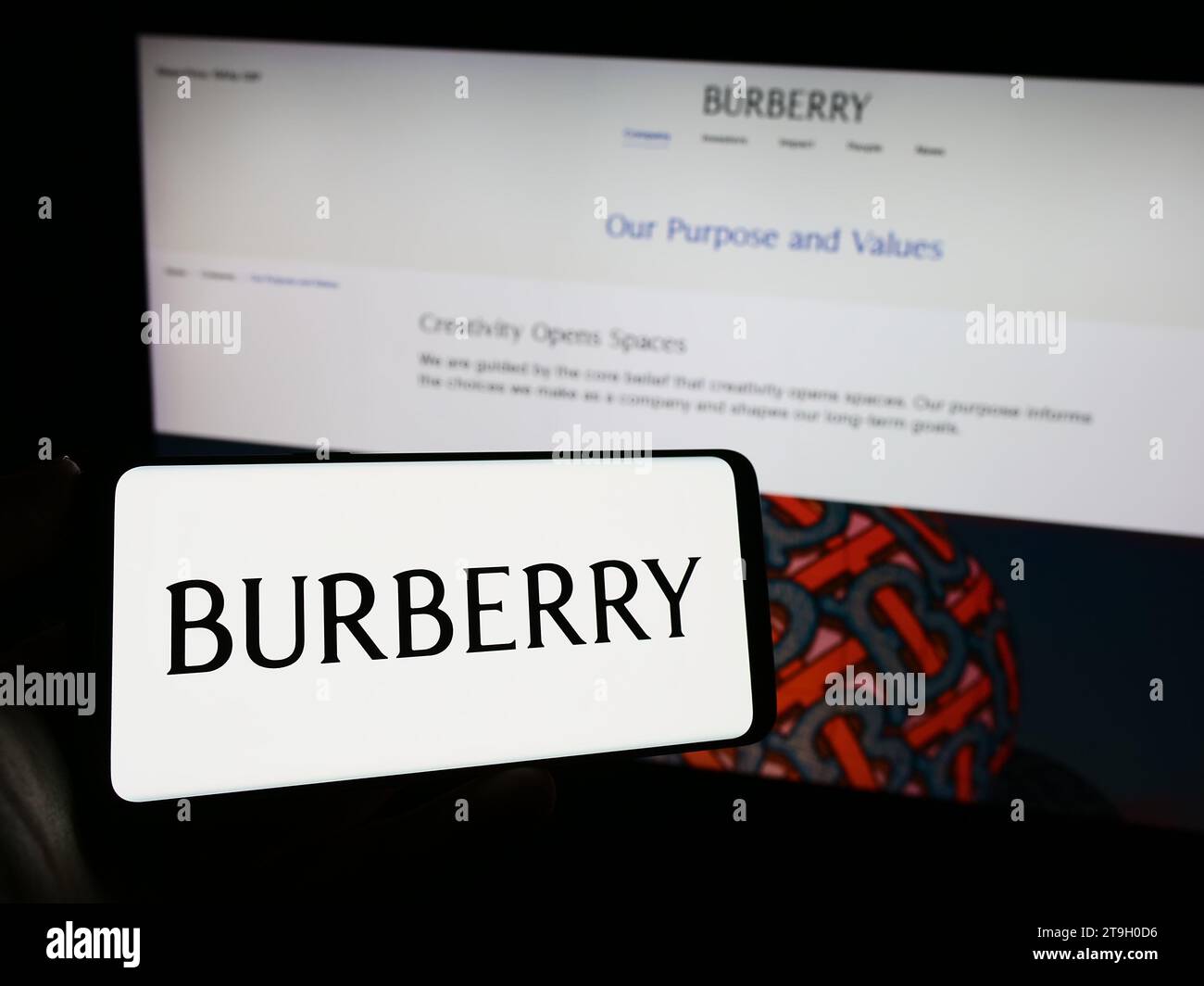 Person, die ein Mobiltelefon mit dem Logo des britischen Luxusmodeunternehmens Burberry Group plc vor der Business-Webseite hält. Schwerpunkt auf dem Display des Telefons. Stockfoto