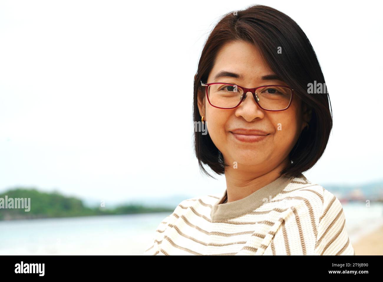 Porträt einer asiatischen Frau mittleren Alters mit Brille, lächelnd, Nahaufnahme Porträt, draußen mit Himmel und Meer Hintergrund. Stockfoto