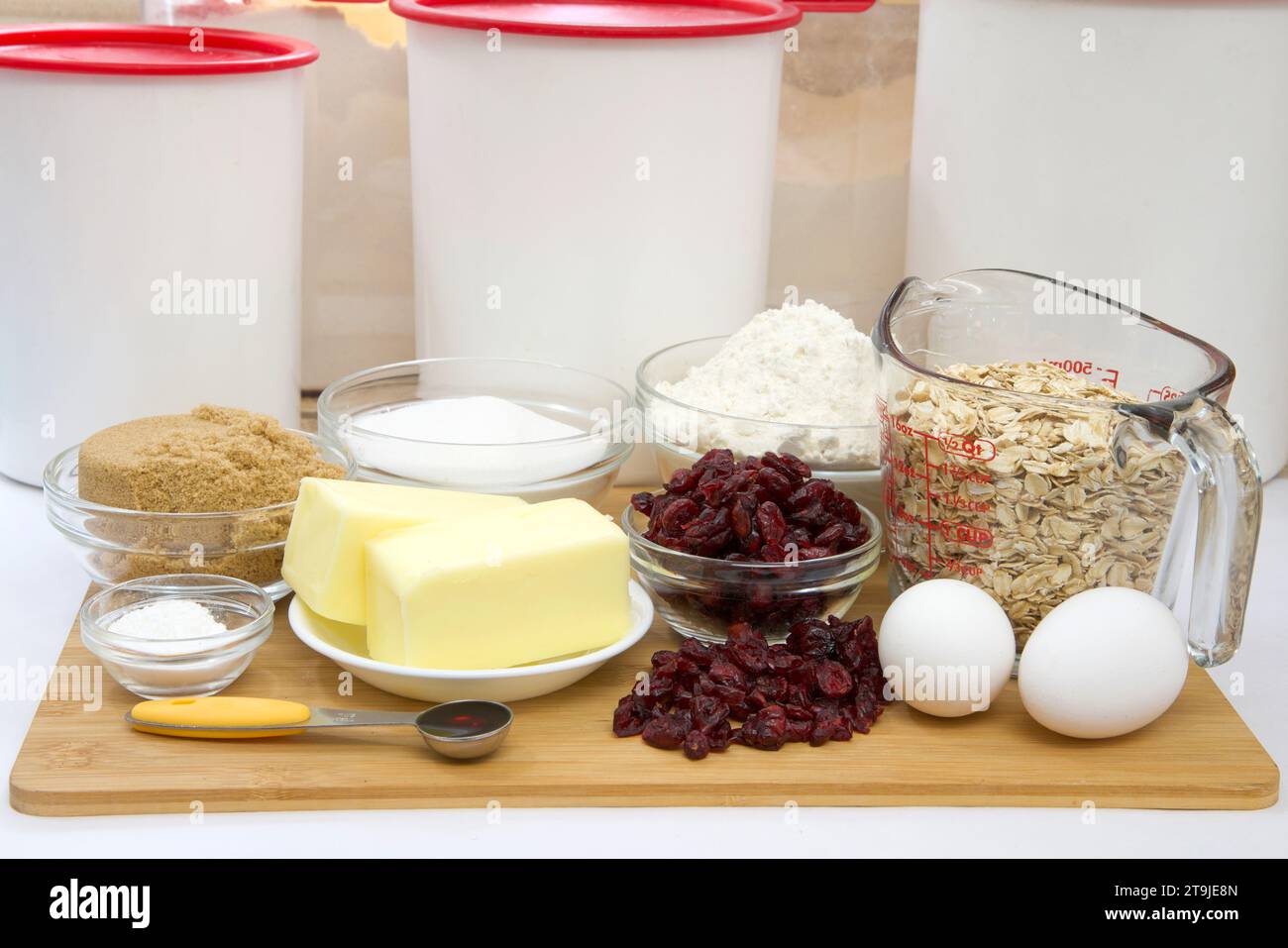 Gemessene Zutaten in Schüsseln und Bechern für Cranberry Haferflockenkekse. Auf Holzschneidebrett mit Pergamentpapier darunter. Kunststoffkanister im Backg Stockfoto
