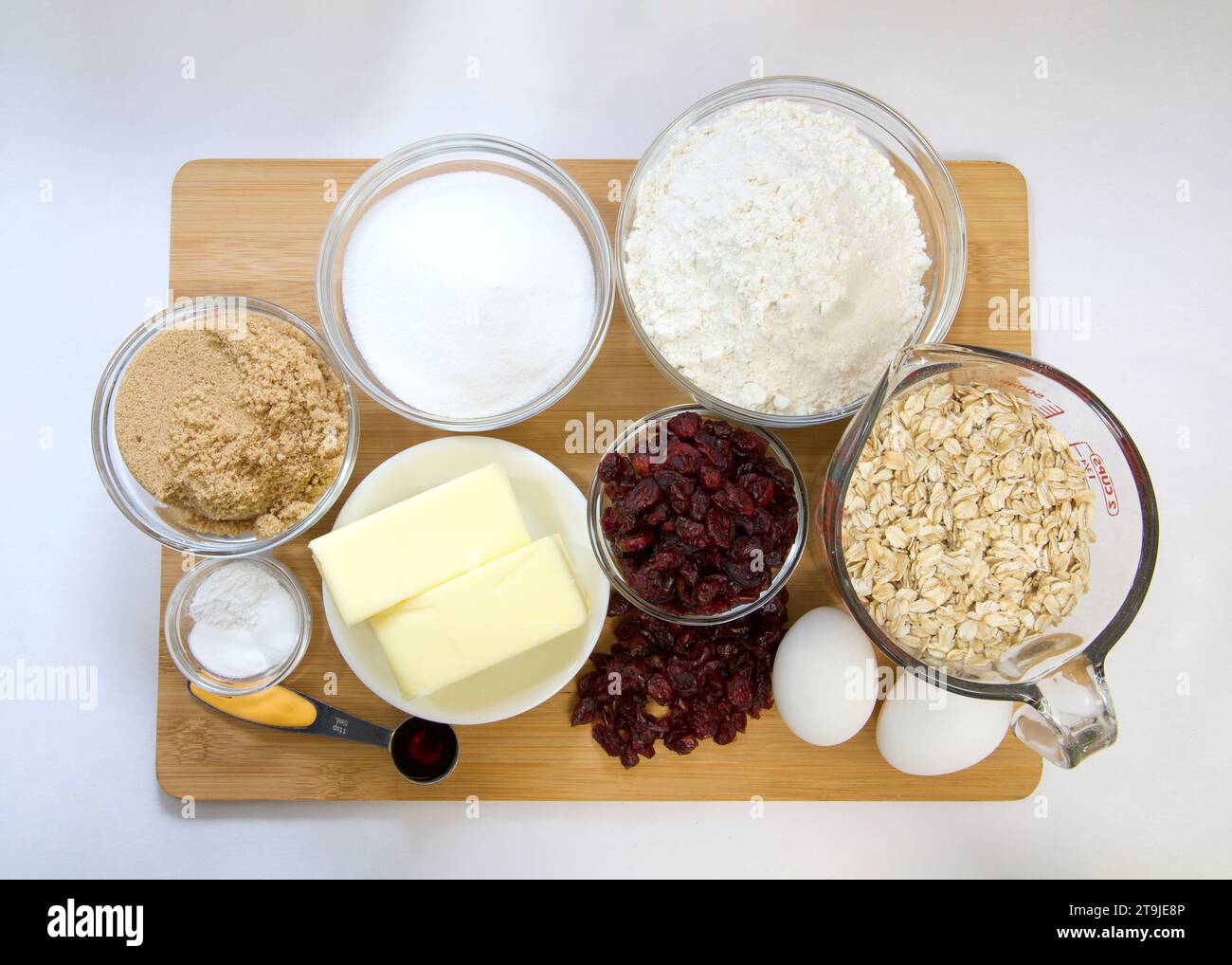 Flache Anordnung der abgemessenen Zutaten in Schüsseln und Bechern für Cranberry-Haferflockenkekse. Auf Holzschneidebrett mit Pergamentpapier darunter. Stockfoto