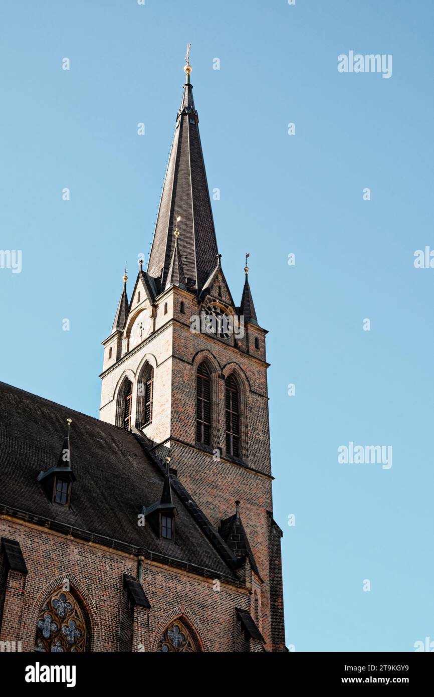 Das Bild zeigt eine Backsteinkirche mit einem hohen Turm und einer Uhr auf dem Turm. Die Kirche hat gewölbte Fenster und wird von der Sonne vor einem blauen Himmel beleuchtet. S Stockfoto