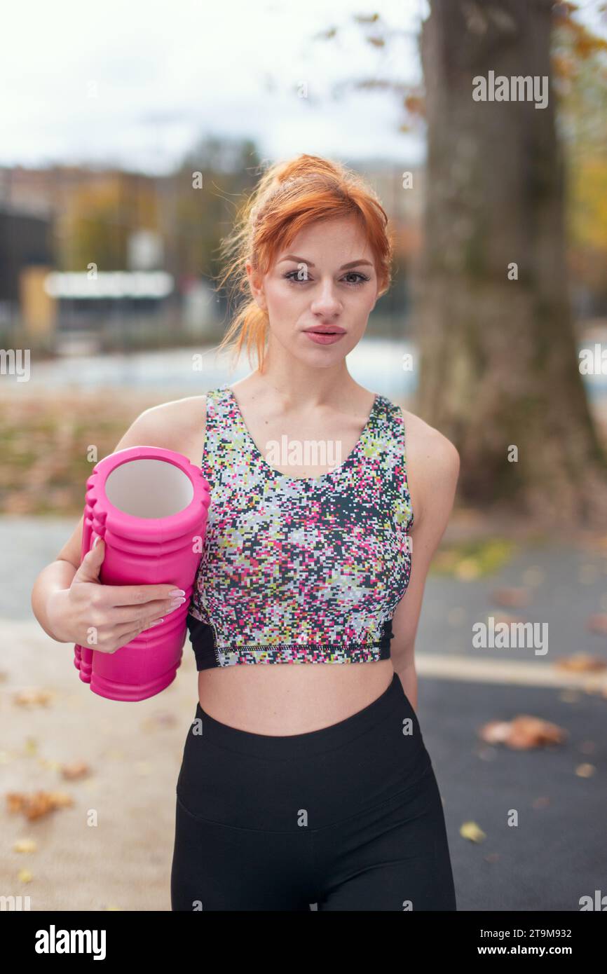Junge rothaarige, selbstbewusste kaukasische Sportlerin, die im Herbst im Park eine Schaumwalze hält Stockfoto