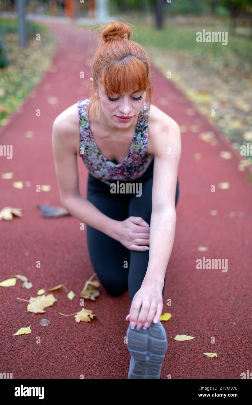 Junge Rothaarige Frau dehnt sich warm, bevor sie im Herbst im Park auf der Rennstrecke läuft Stockfoto