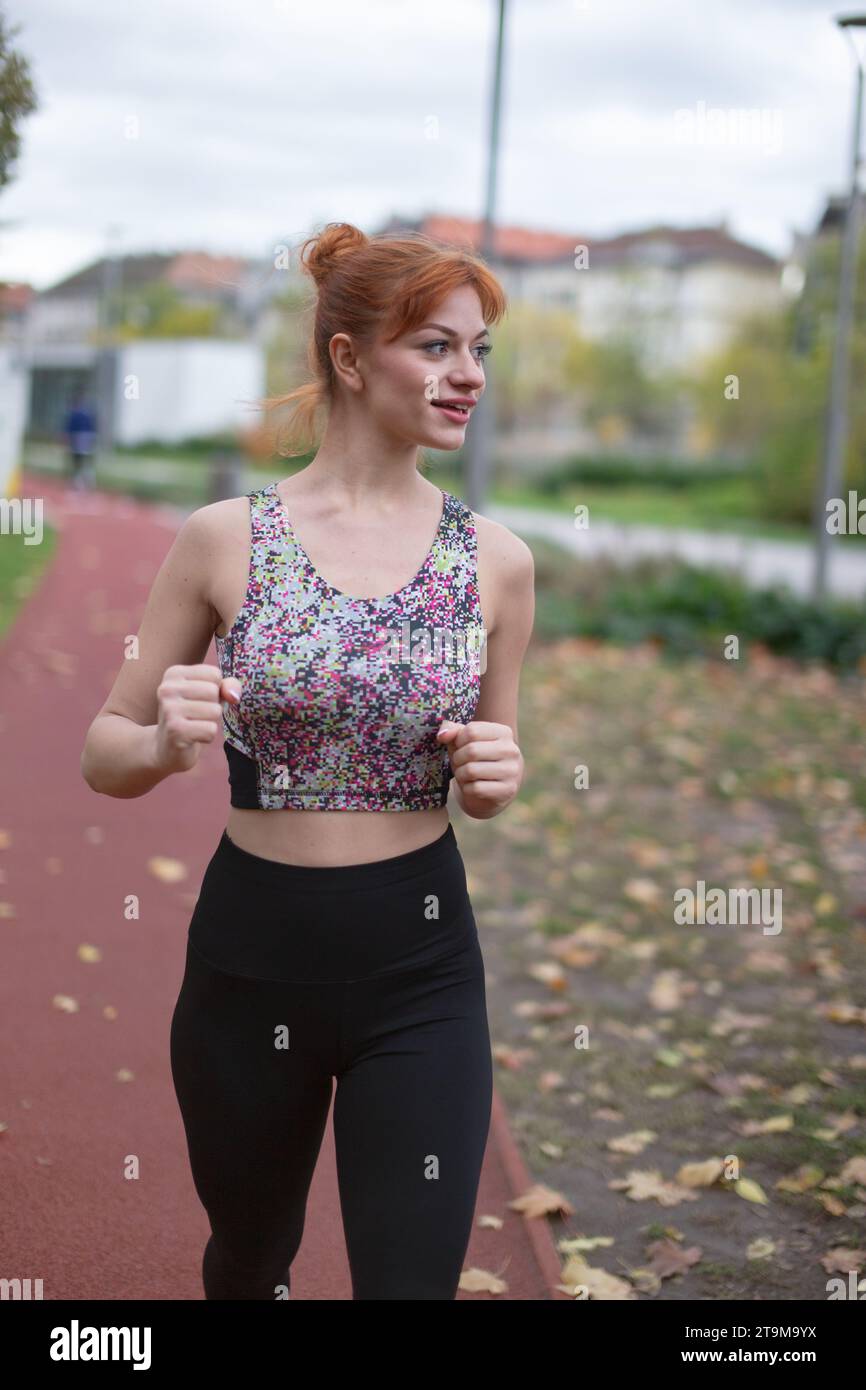 Junge rothaarige weiße Frau in Sportkleidung, die im Spätherbst auf der Laufstrecke joggt und wegblickt Stockfoto