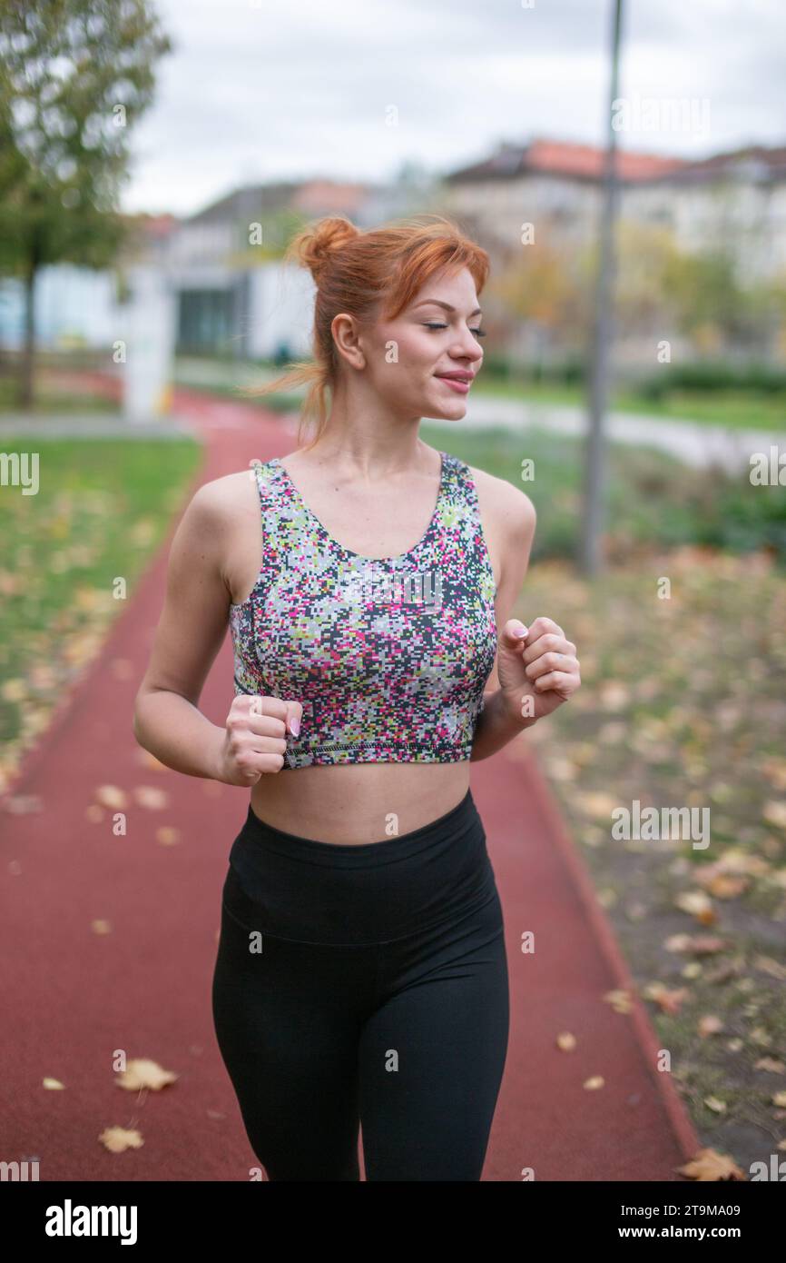 Junge rothaarige, selbstbewusste Kaukasierin in Sportswear, die im Spätherbst auf der Laufbahn joggt, die Augen geschlossen Stockfoto