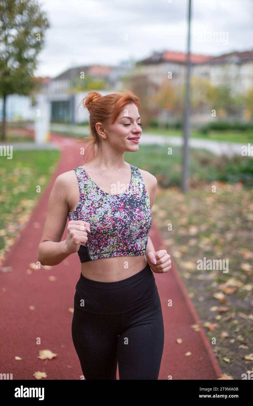 Junge rothaarige positive Kaukasierin in Sportswear, die im Spätherbst auf der Laufstrecke joggt Stockfoto