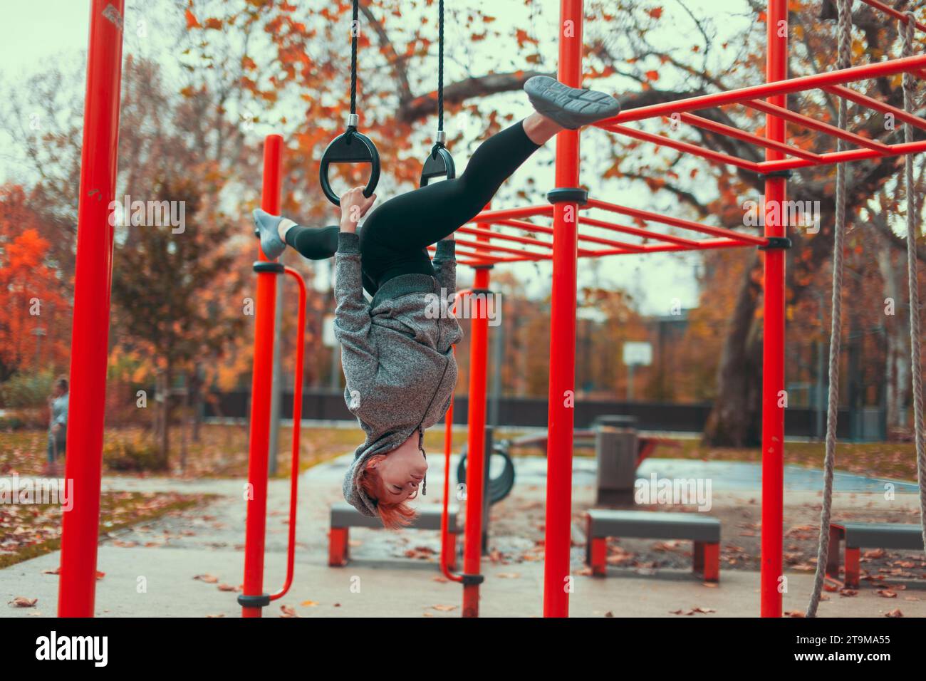 Junge Turnerin in der Stadt Kaukasierin auf dem Spielplatz, die kopfüber an Ringen hängt und im Herbst im Park Spaltungen macht Stockfoto