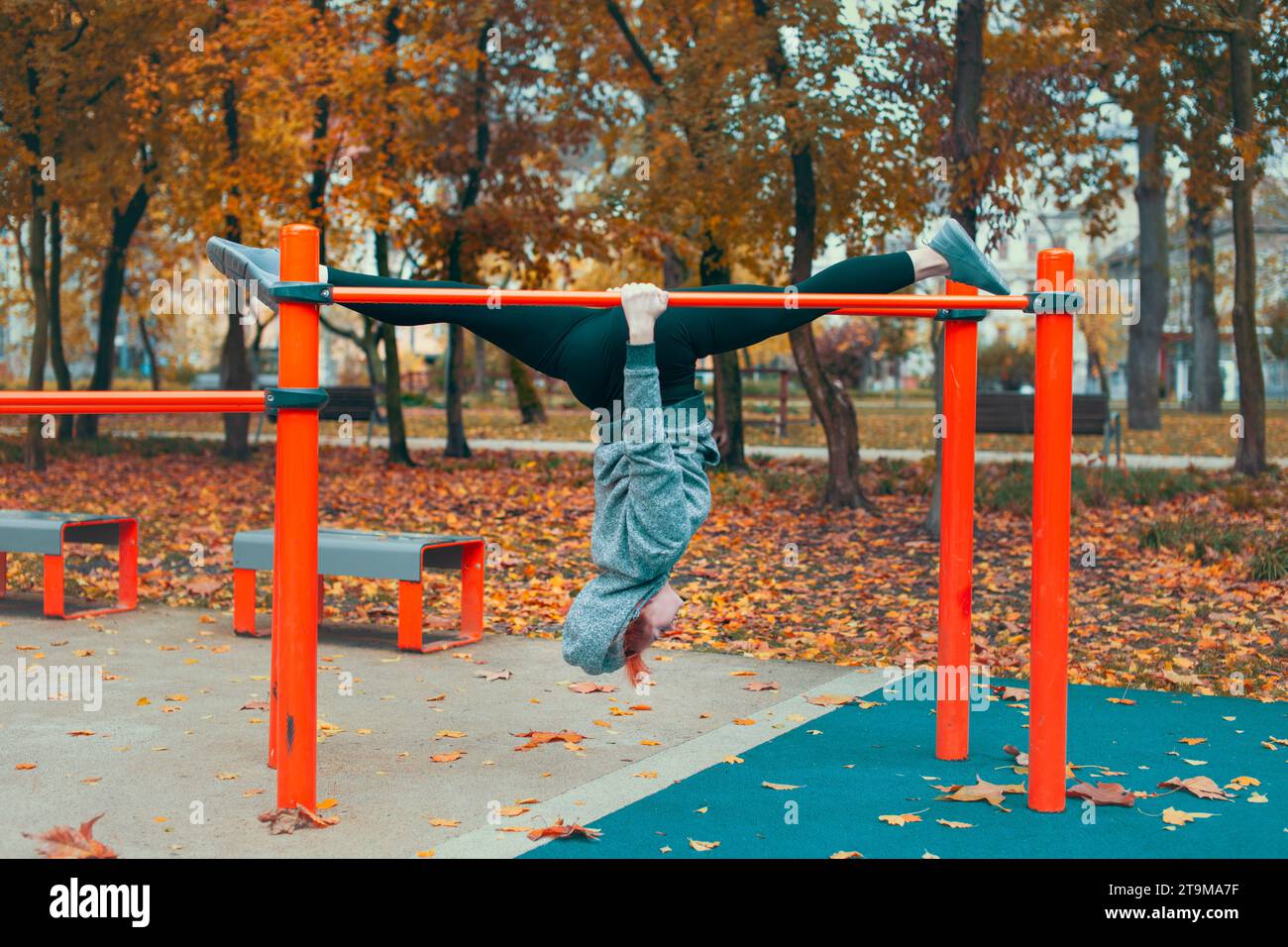 Junge Turnerin in der Stadt Kaukasierin auf dem Spielplatz, die kopfüber an Bars hängt und im Herbst im Park Spaltungen macht Stockfoto