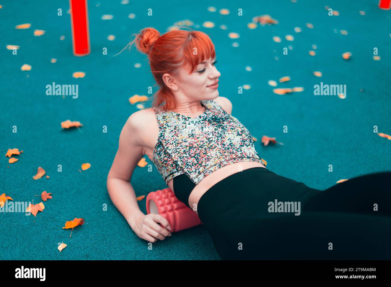 Junge rothaarige Kaukasierin in Sportswear, die sich mit einem Schaumstoffroller aufwärmt, bevor sie im Herbst im Park trainieren Stockfoto