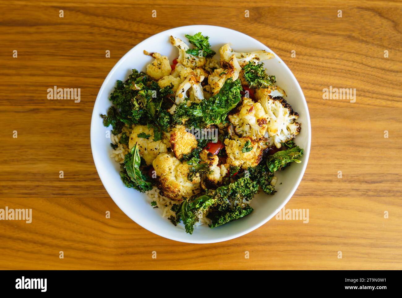 Vegetarische Mahlzeit mit im Ofen geröstetem Blumenkohl, Grünkohl und roter Paprika über braunem Reis in einer weißen Schüssel auf einem Holztisch. Stockfoto
