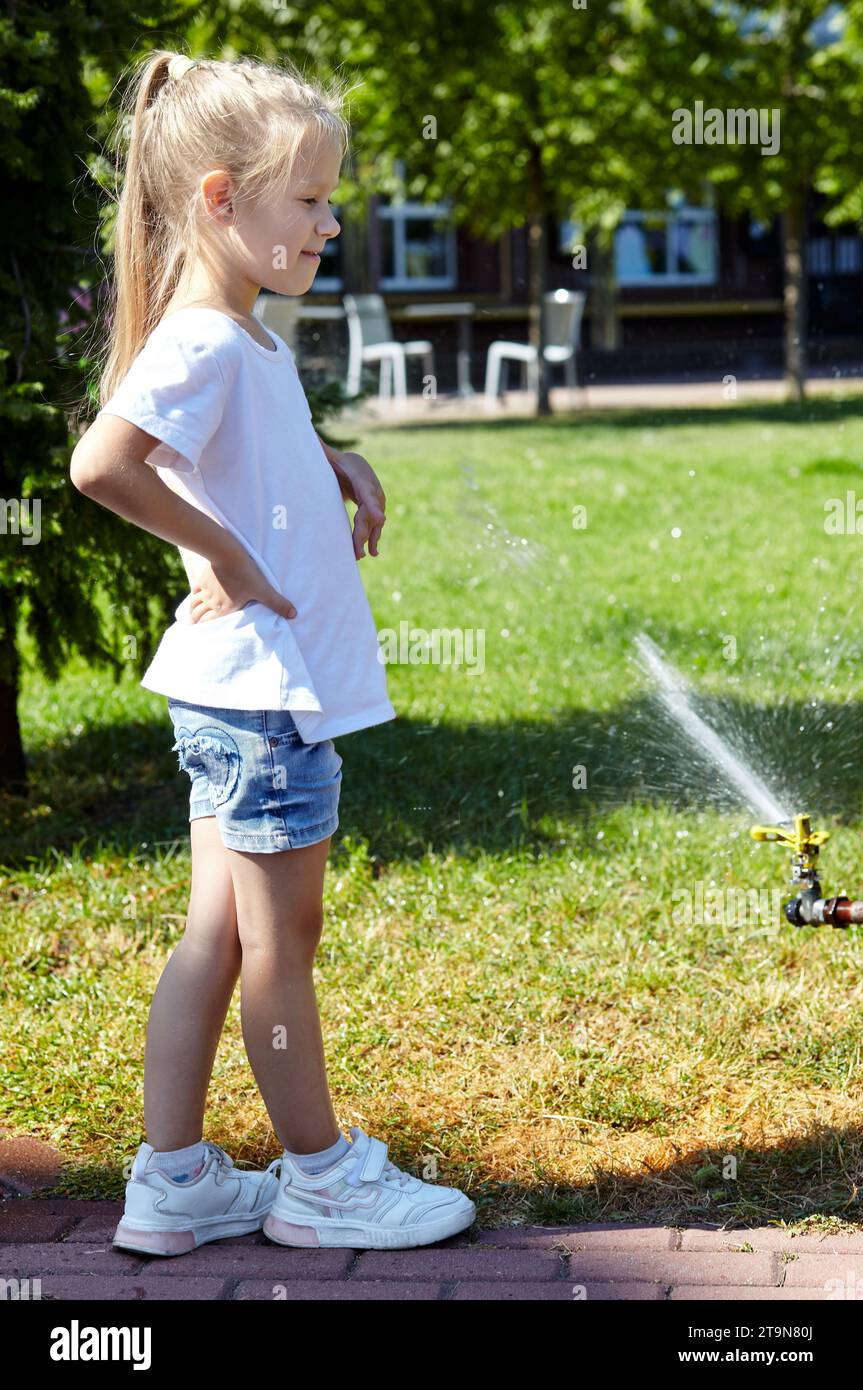Kleines Mädchen, das unter dem Wasser von der Sprinklerbewässerung im Sommer Stadtpark läuft. Kindheit, Freizeit und Menschenkonzept - glückliche Kinderruhe und Stockfoto