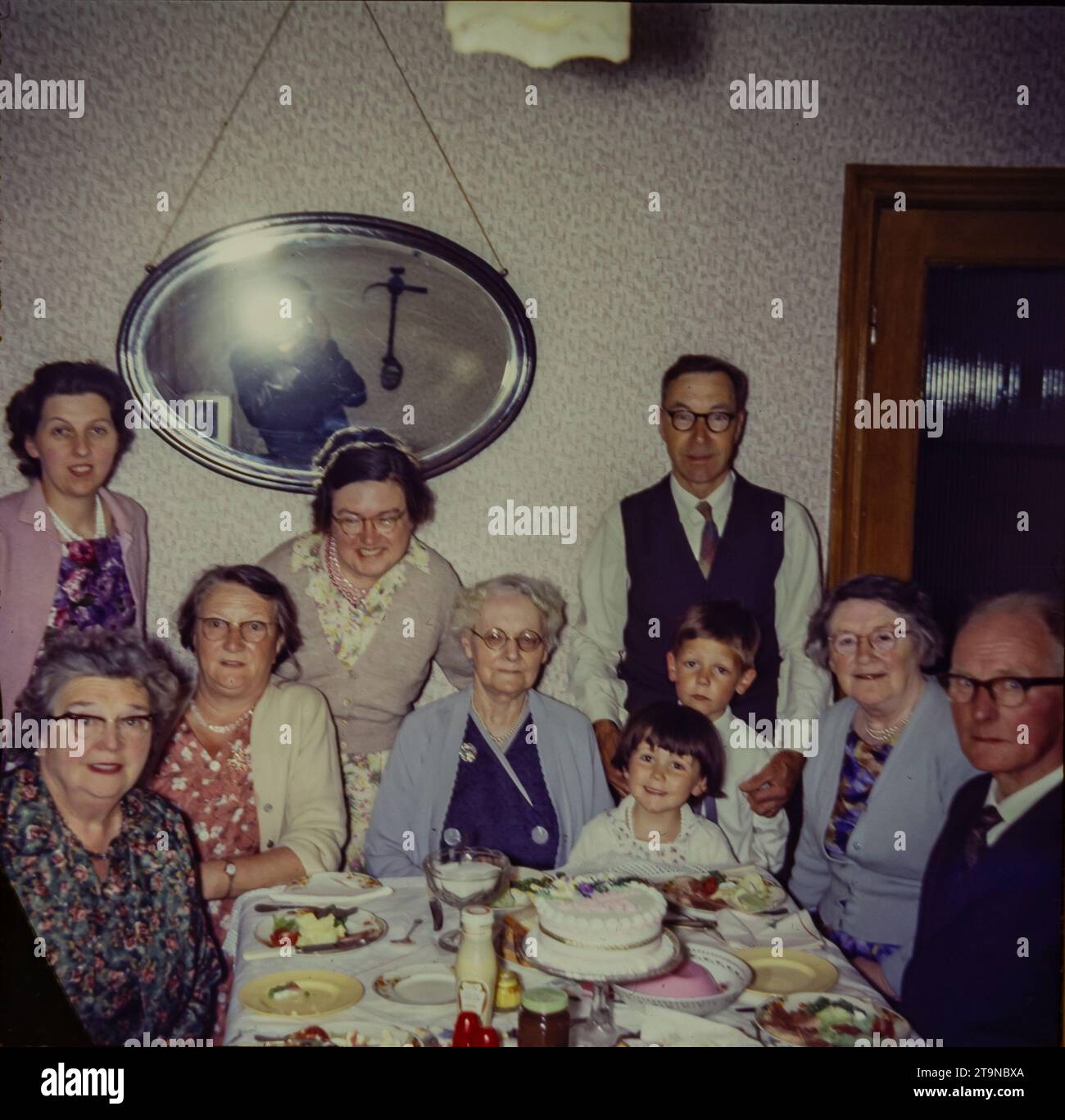 Eine Großfamilie, die 1962 Geburtstag feiert. Es gibt vier Generationen von Familien, die um einen Tisch sitzen. Das Foto zeigt einen Speisesaal aus den 1960er Jahren, Mode aus den 1960er Jahren und einen Nachmittagstee mit Salat, Blancmange und Kuchen. Dies ist ein Foto, das von der Originalfolie aufgenommen wurde. Stockfoto