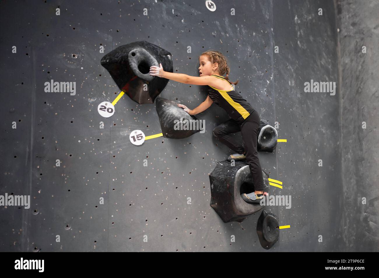 Das kleine Mädchen trainierte klettert in der Kletterhalle an der Wand. Kind mit braunem Haar in Sportkleidung, das ohne Hilfe im Boulderzentrum für Kinder arbeitet. Das tapfere und starke Mädchen klettert auf die falsche Wand Stockfoto