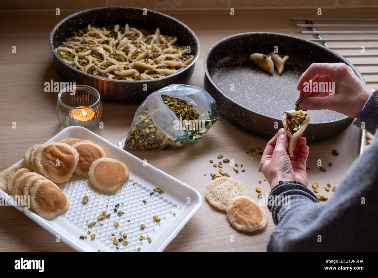 Hände halten Qatayef gefüllt mit Schokolade und mit Pistazien belegt auf einem Holztisch, mit einem Teller, der später im Ofen als Ramadan swe zubereitet wird Stockfoto