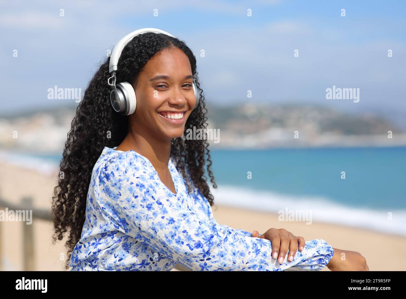 Glückliche schwarze Frau, die Kopfhörer trägt, schaut dich an einem sonnigen Tag am Strand an Stockfoto