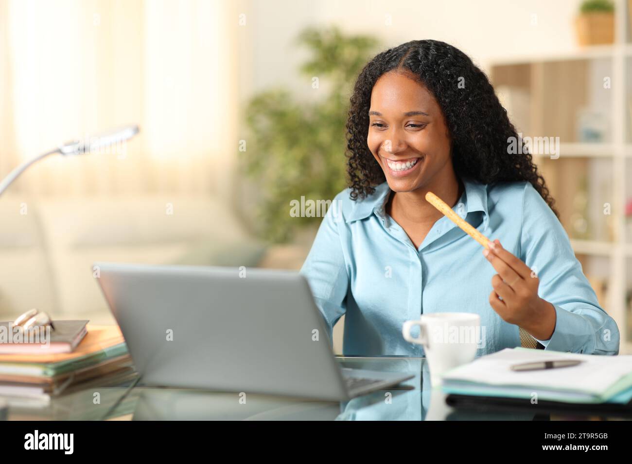 Glücklicher schwarzer Tele-Mitarbeiter, der zu Hause einen Snack isst und mit einem Laptop arbeitet Stockfoto