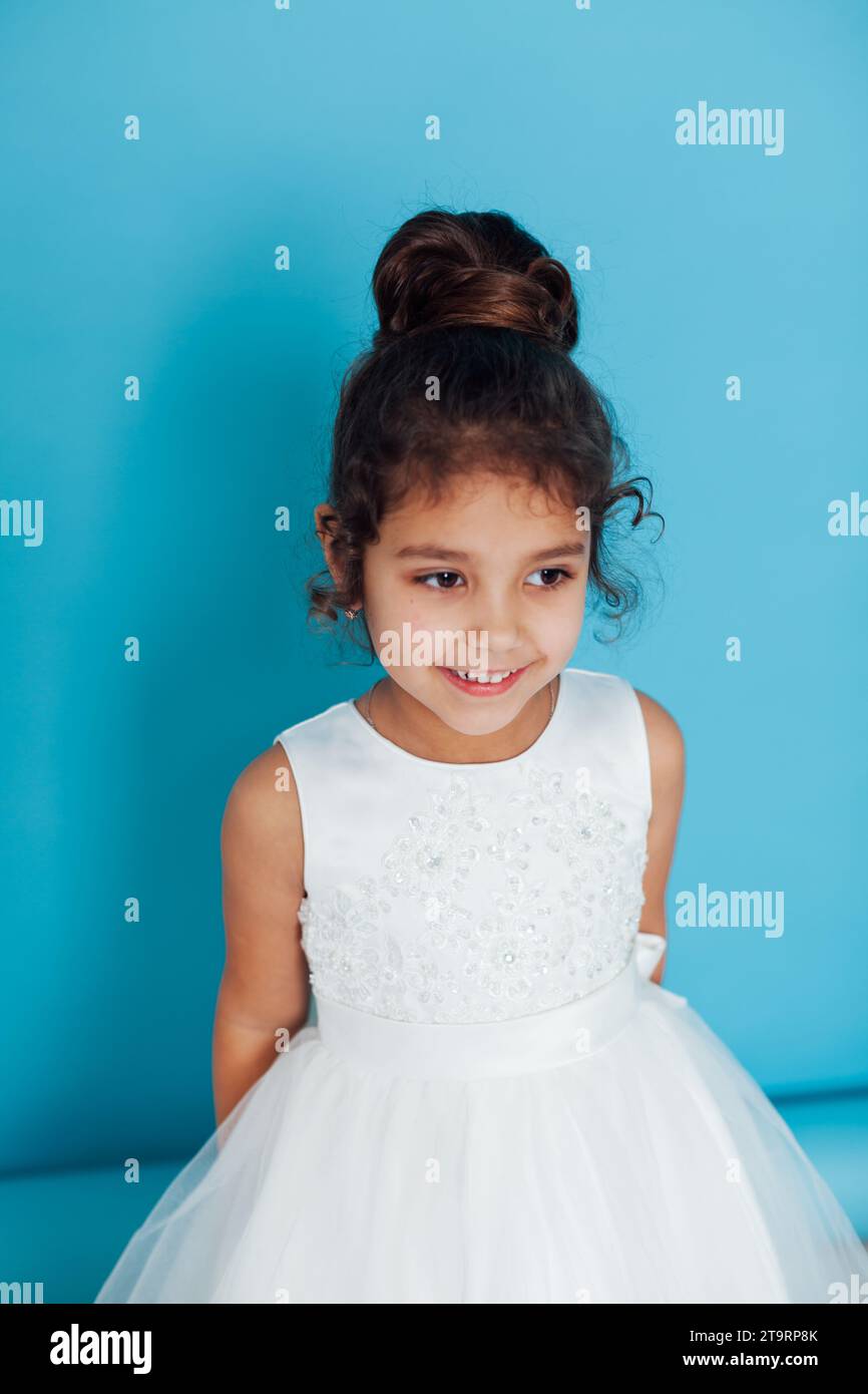 Ein kleines Mädchen in einem weißen Kleid mit Frisur posiert auf blauem Hintergrund Stockfoto