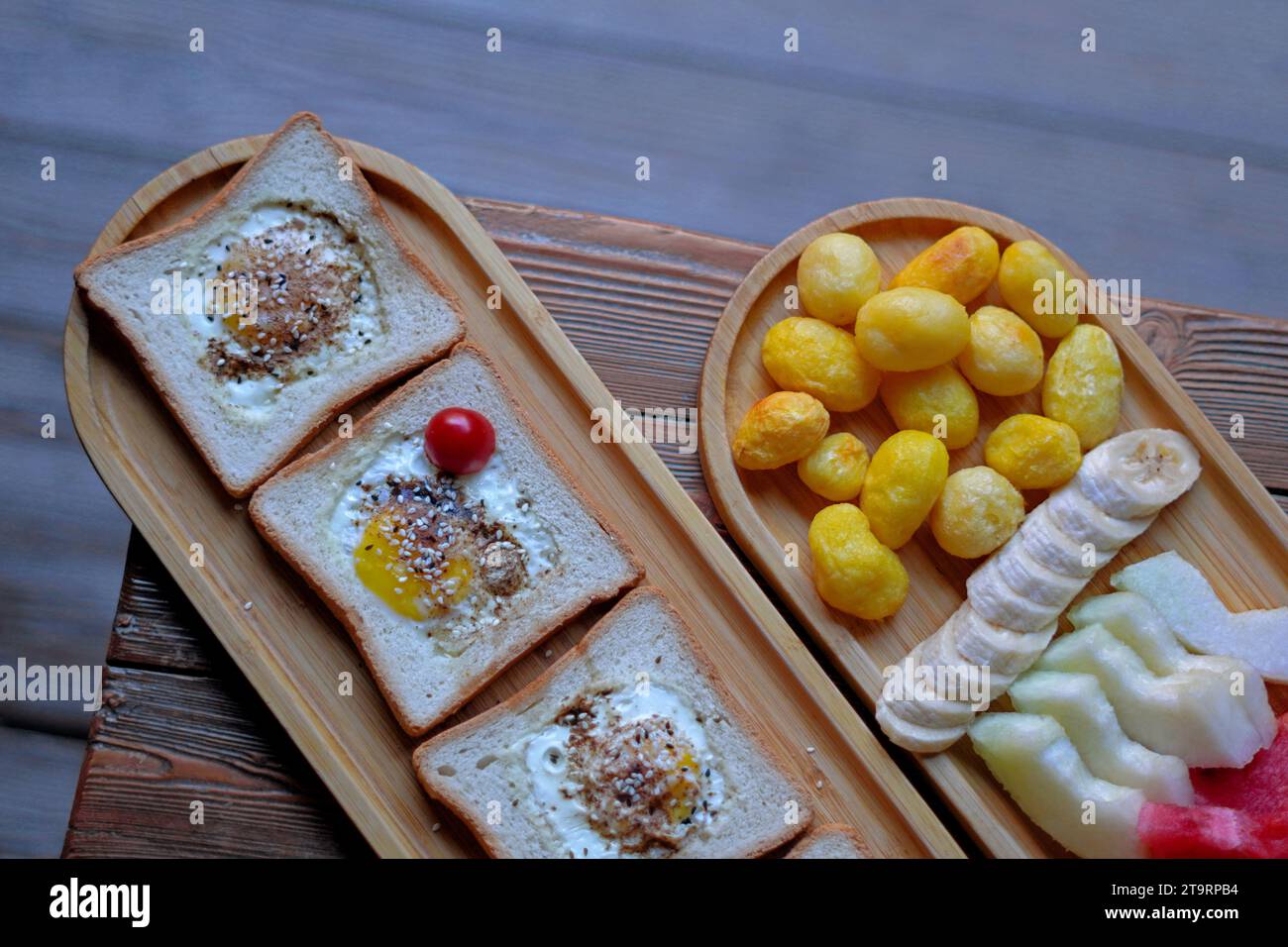 Ein Teller mit geröstetem Brot und Eiern, neben einem Teller mit gebackenen Kartoffeln, geschnittenen Bananen, Melonen und Wassermelonen. Stockfoto