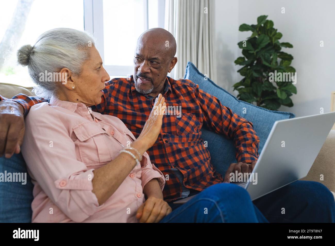 Vielseitiges Seniorenpaar, das auf dem Sofa sitzt und im sonnigen Wohnzimmer einen Laptop nutzt. Lebensstil, Ruhestand, Seniorenleben, Zweisamkeit, Kommunik Stockfoto
