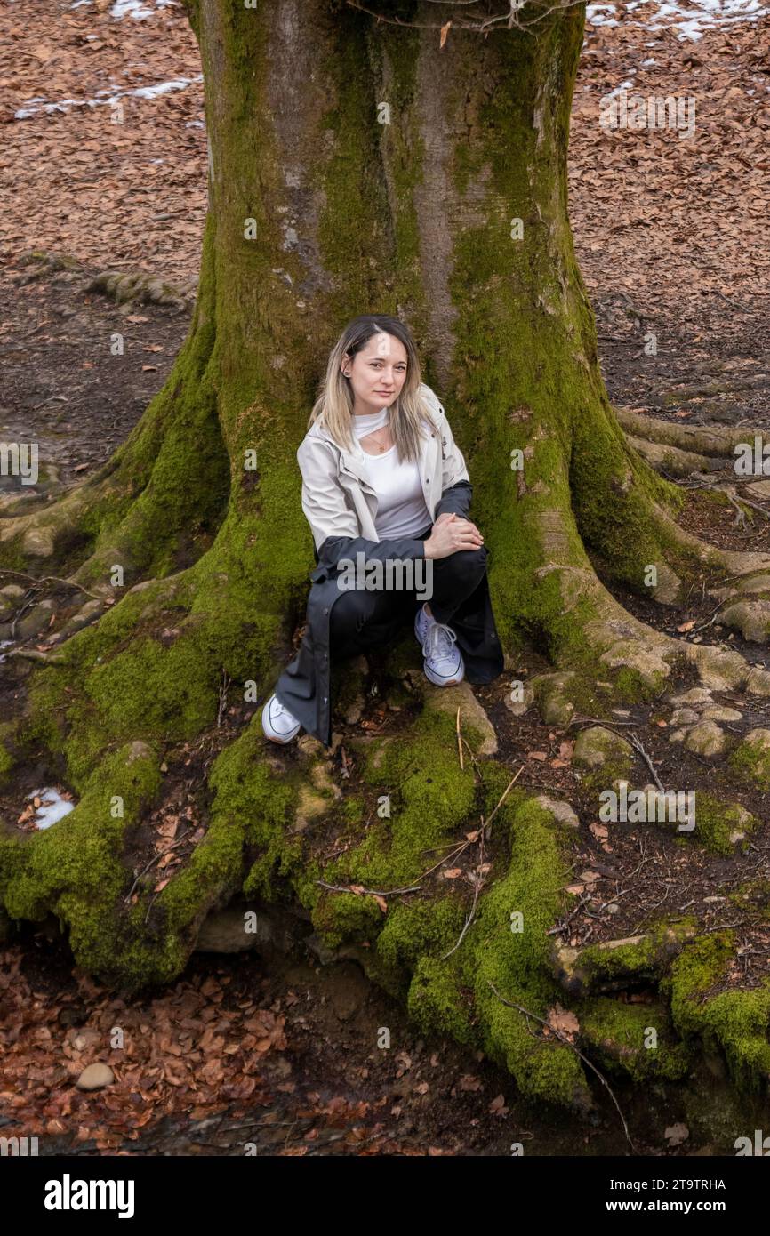 Eine Frau in weißem Hoodie und schwarzer Hose sitzt auf den moosbedeckten Wurzeln eines Baumes in einem Wald, neben einem kleinen Bach Stockfoto