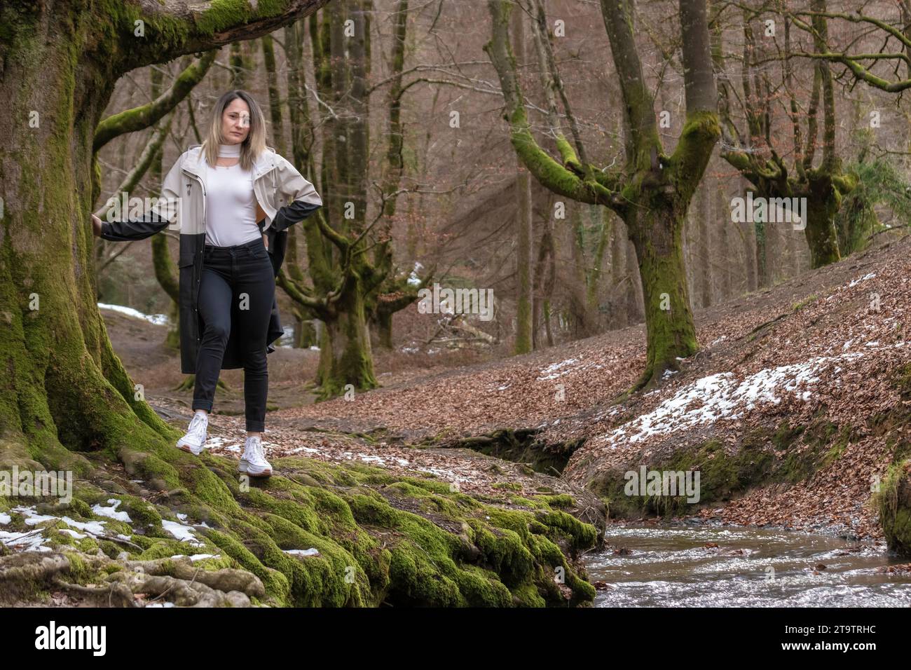 Frau in weißem Hemd und schwarzer Hose steht auf einem moosigen Felsen in einem Wald, Arme ausgestreckt, mit einem Bach, der durch die Mitte läuft Stockfoto