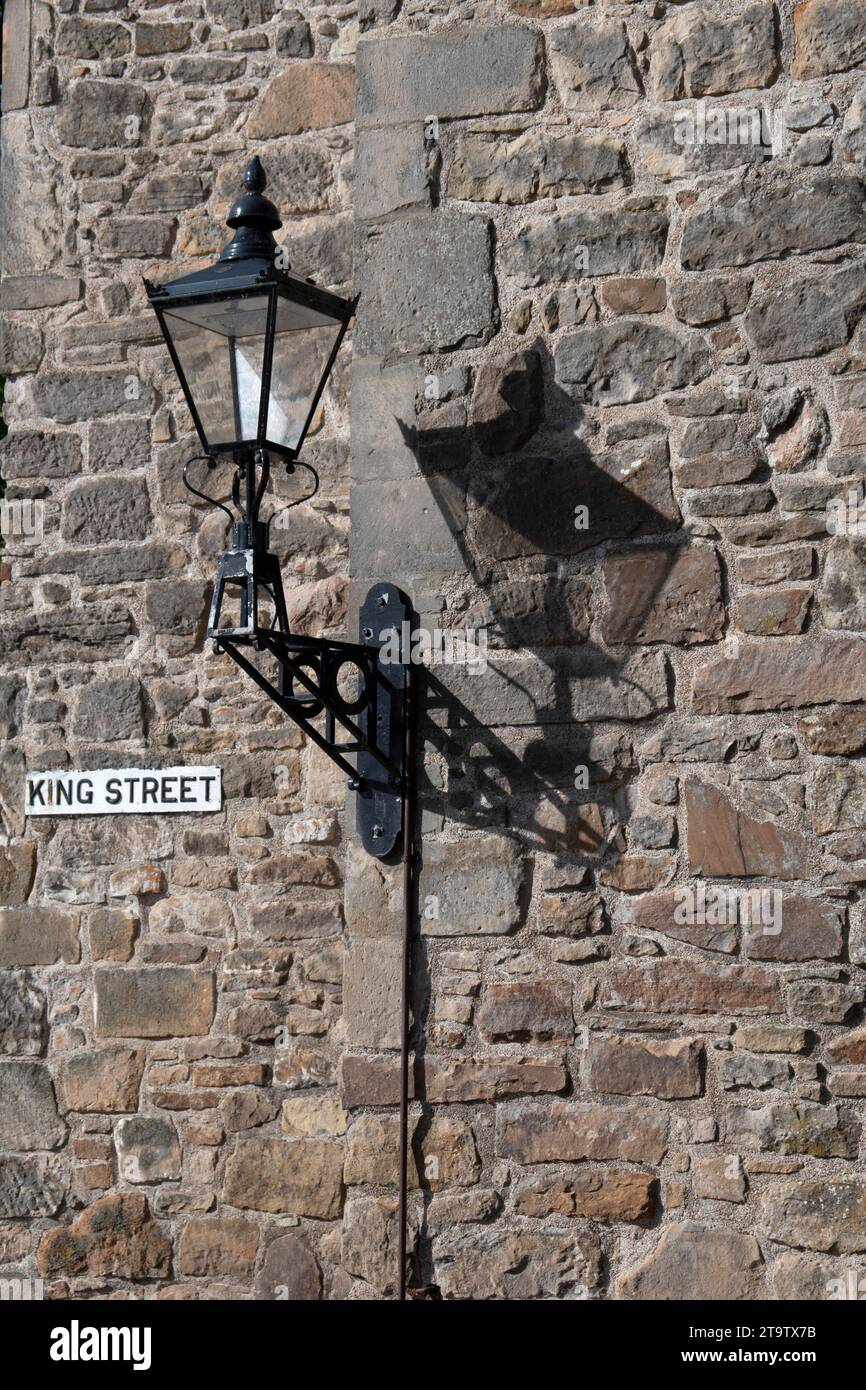 Elektrisches Licht im alten Laternen-Stil, das Schatten von der Sonne auf eine Steinmauer wirft. Stockfoto