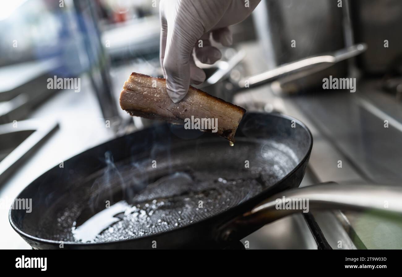 Die Hand hält einen knusprigen Schweinebauch, der in einer heißen, geölten Pfanne an einem Gasherd in einer professionellen Küche eines Restaurants gebraten wurde. Küche im Luxushotel Stockfoto