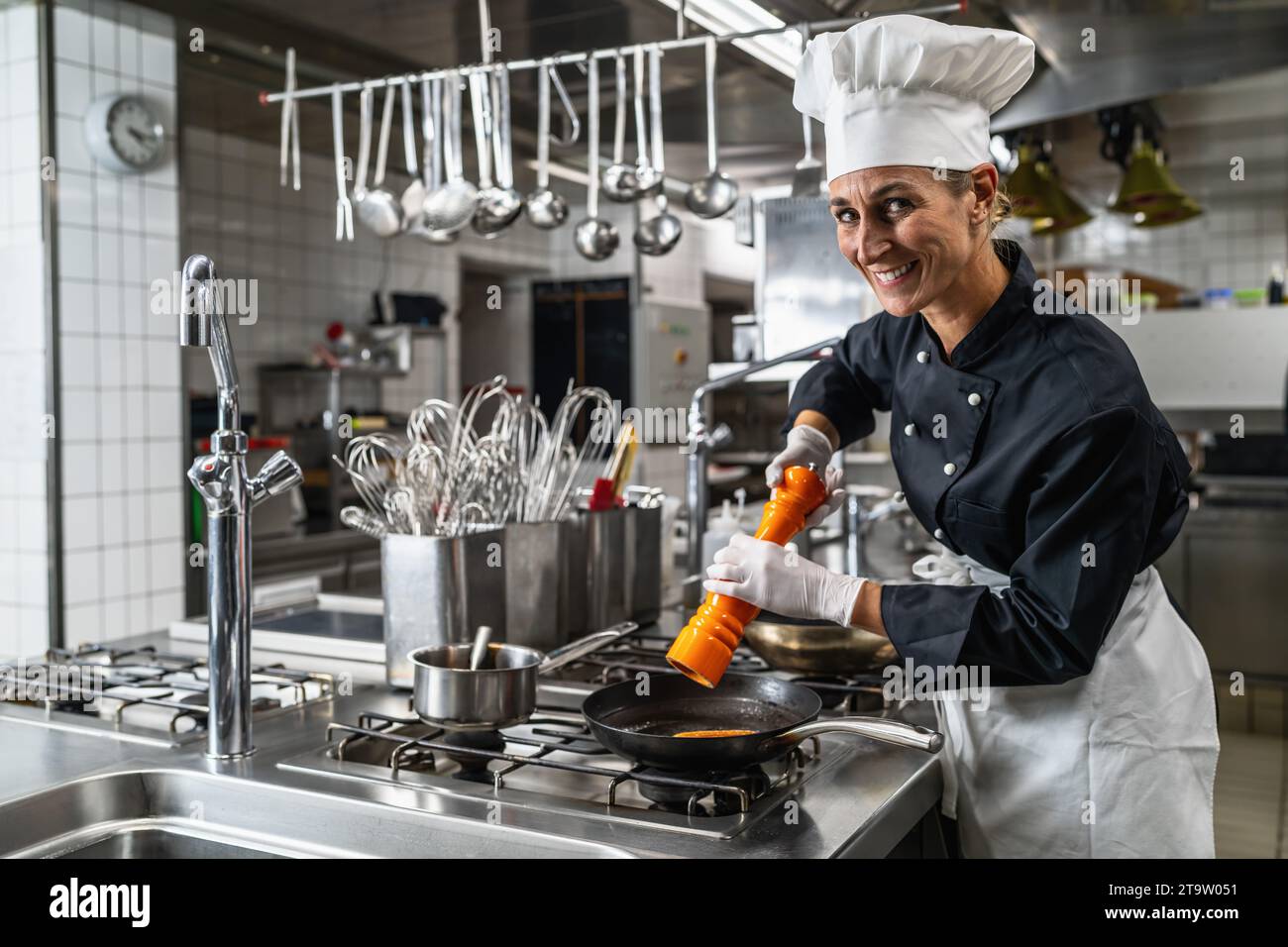 Lächelnde Küchenchefin in der Hotel- oder Restaurantküche kocht und würzt das Essen mit einer Pfeffer- oder Salzmühle Stockfoto