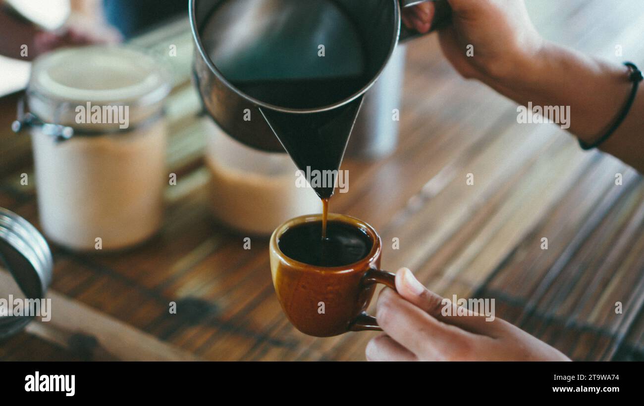 Barista gieße Kaffee in die Tasse auf Tisch, Bambusboden Hintergrund. Mittelteil der Frau, die Kaffee in eine Tasse gießt. Thailändischer lokaler Stil. Stockfoto