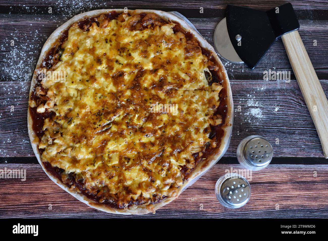 Frisch gebackene, hausgemachte Pizza, serviert auf einem bemehlten Holztisch. Leckere Hühnerpizza mit Käse. Pizzaschneider, Salz- und Pfefferstreuer. Draufsicht. Stockfoto