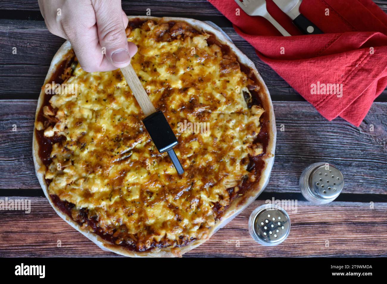 Die Hand des Mannes schneidet hausgemachte Pizza auf Scheiben mit einem Spezialmesser. Hühnchenkäse-Pizza auf Holztisch. Rote Serviette, Messer und Gabel, Salz und Pfeffer. Stockfoto