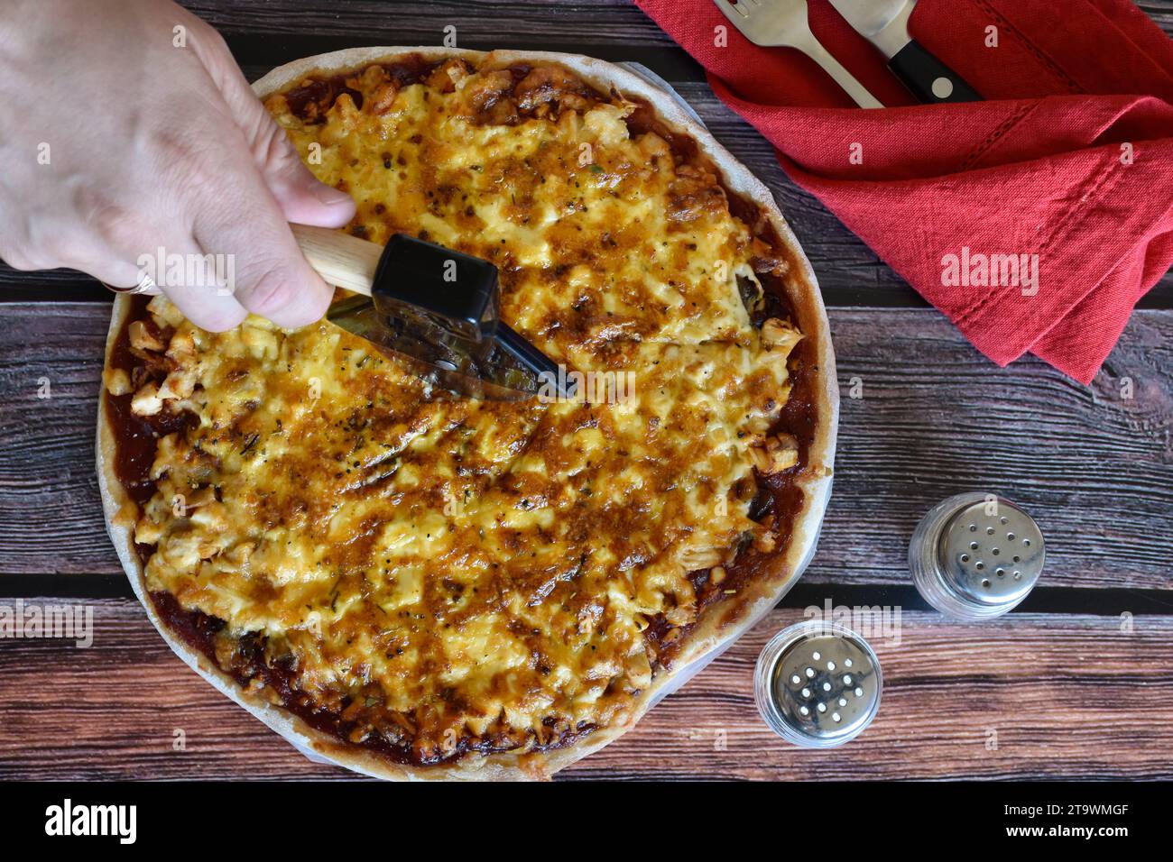 Die Hand des Mannes schneidet hausgemachte Pizza auf Scheiben mit einem Spezialmesser. Hühnchenkäse-Pizza auf Holztisch. Rote Serviette, Messer und Gabel, Salz und Pfeffer. Stockfoto