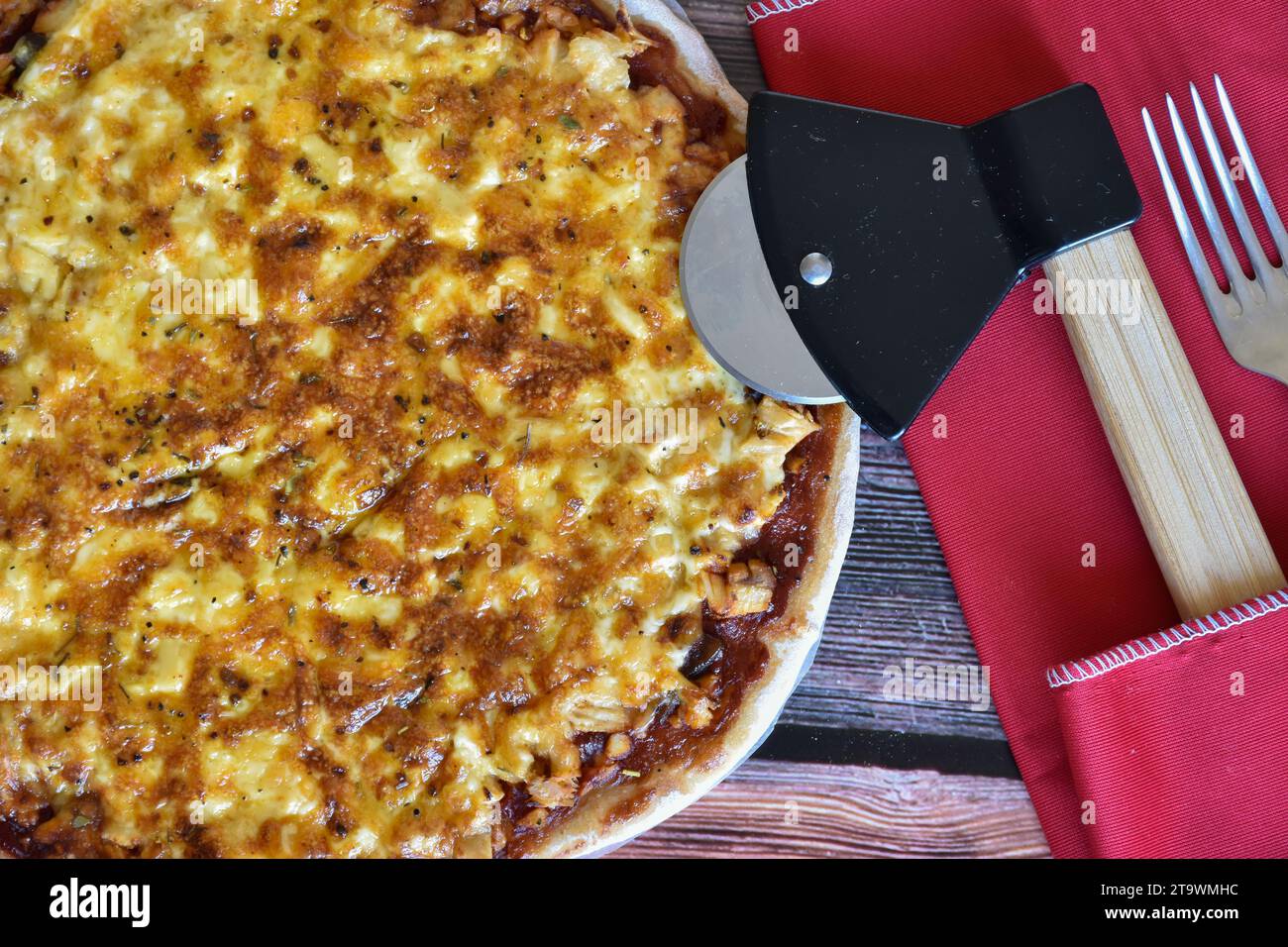 Frisch gebackene hausgemachte Pizza auf rustikalem Holztisch. Leckere Hühnerpizza mit Käse. Pizzaschneider, rote Serviette, Messer und Gabel. Draufsicht. Stockfoto