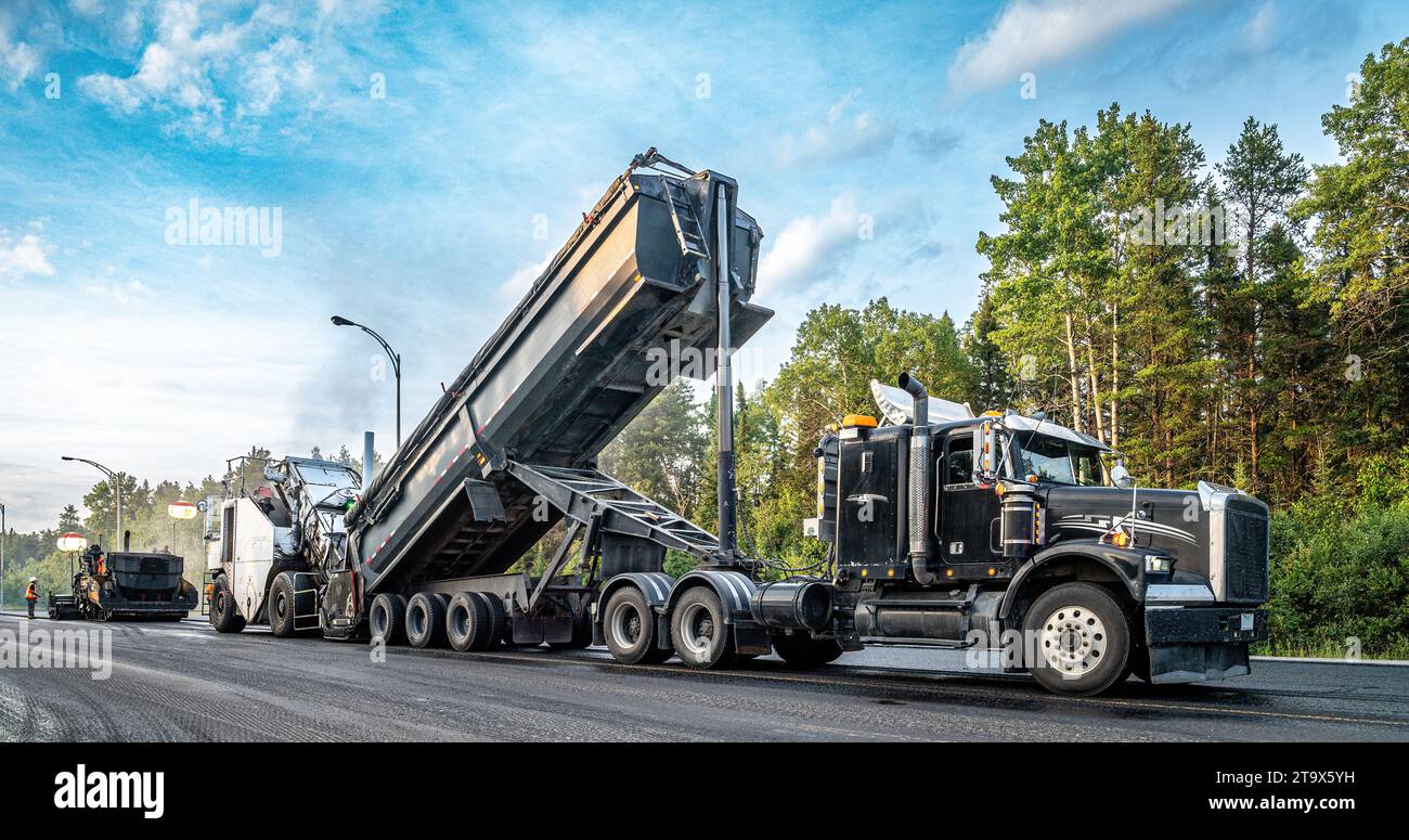 Sattelzugwagen entladen Asphalt in ein Transportfahrzeug während der Pflasterarbeiten auf dem Trans-Canada Highway, spät am Tag Stockfoto