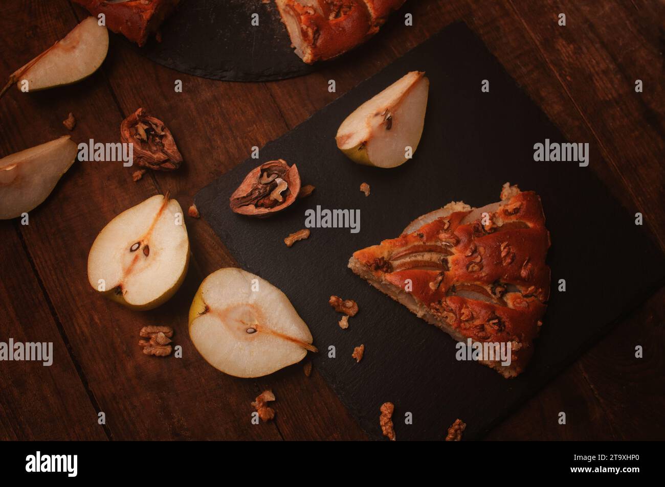 Birnenkuchen auf einem schwarzen Servierbrett auf einem Holztisch, geschnittene Birnen und Walnüsse liegen chaotisch Stockfoto