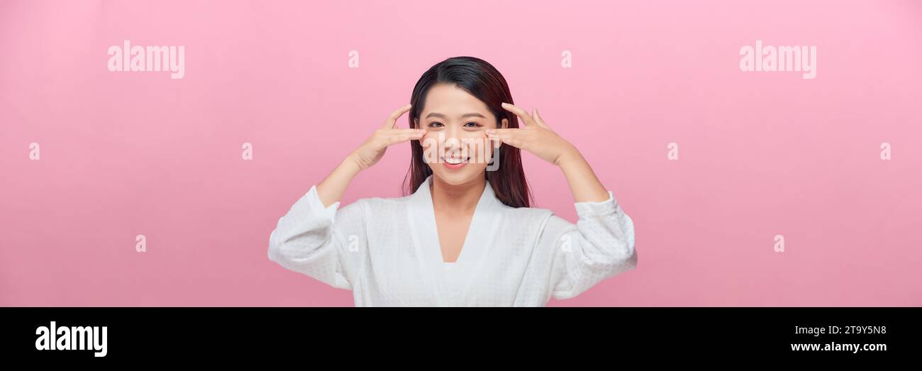 Schönes lächelndes asiatisches weibliches Modell mit natürlichem Make-up, das glühende, hydrierte Haut auf rosa Hintergrund berührt. Stockfoto